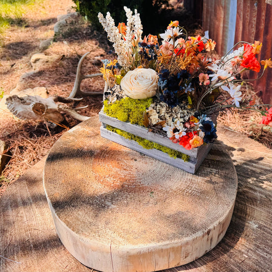 A rustic flower arrangement in a weathered gray wooden crate lined with bright green moss, displayed on a round natural wood log slice outdoors.